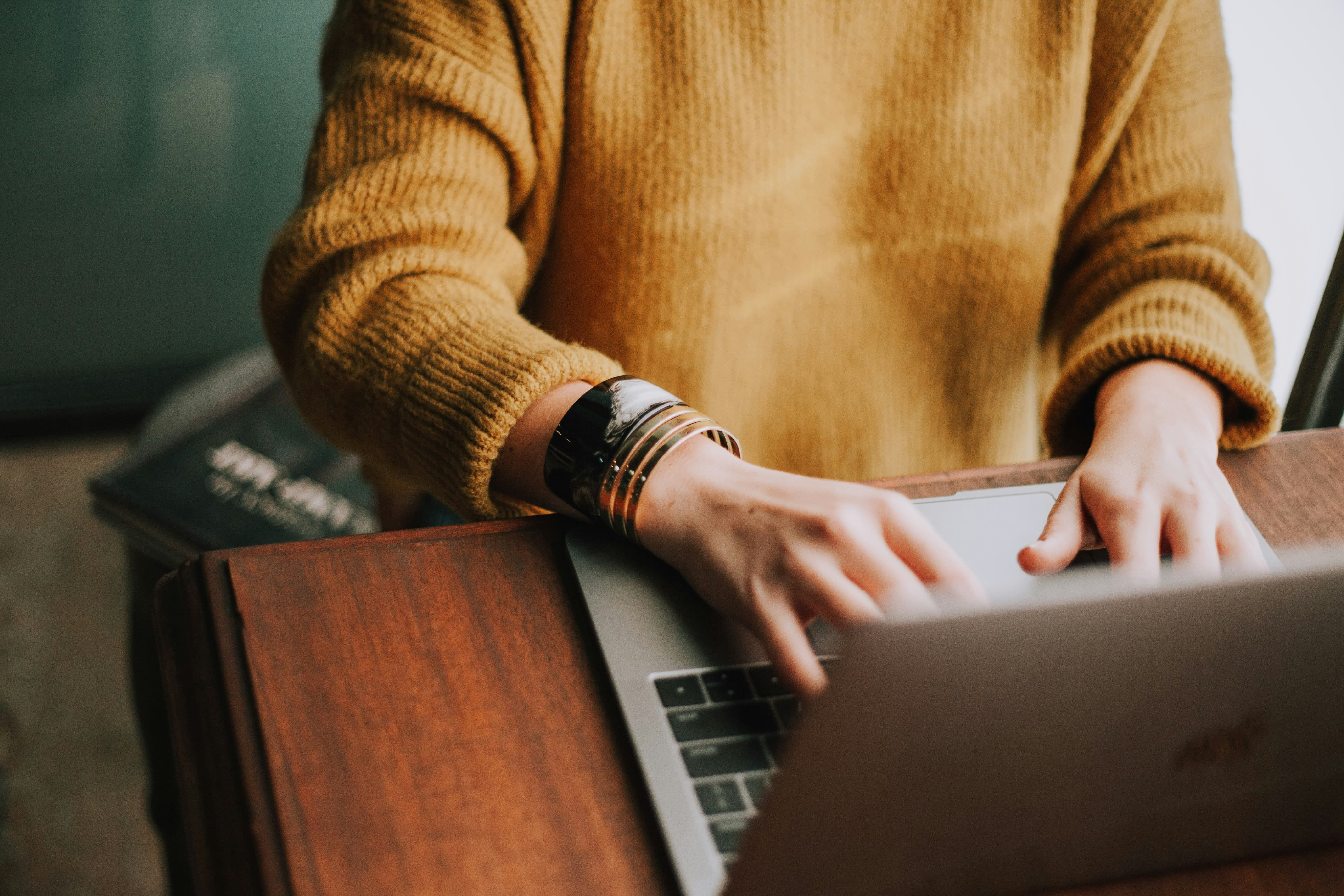 A person wearing an orange sweater and bangles typing at a computer. Their face is not visible.